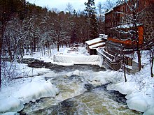 Paysage de neige. Au premier plan, cascade d'un petit cours d'eau, près duquel s'alignent deux bâtiments.