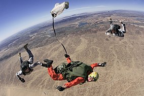 Студент спецназа с большой высотой и низким уровнем подготовки тянет за разрывной шнур, в то время как два инструктора наблюдают и критикуют его прыжок на полигоне Юма, штат Аризона.