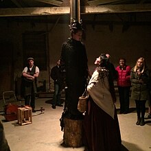 warm dim lighting illuminates two women actors in period costumes (long skirts), one is stading on a tree stump in front of a post, the other is looking up to her. A few in audience stand around the side