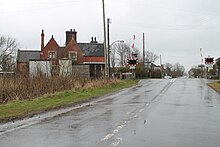 On a wet day, with rain falling, we are standing around 400 yards from a level crossing on a wide but not busy road. The crossing, protected by lifting barriers, is next to the former station, a building in red brick with stone Mullions.