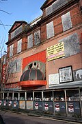 Boarded up frontage of a red-brick building, with two porticoes on the top floor, a partially-melted semicircular shelter over the ground floor, and a boarded walkway obscuring the ground floor entrance.