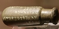 A bronze bottle on its side with text in the Phoenician alphabet "𐤅𐤀𐤔𐤇𐤕 / 𐤉𐤂𐤋 / 𐤅𐤉𐤔𐤌𐤇 /𐤁𐤉𐤅𐤌𐤕 𐤓𐤁𐤌 𐤅𐤁𐤔𐤍𐤕"