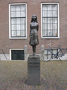 A bronze statue of a smiling Anne Frank, wearing a short dress and standing with her arms behind her back, sits upon a stone plinth with a plaque reading "Anne Frank 1929–1945".  The statue is in a small square, and behind it is a brick building with two large window, and a bicycle.  The statue stands between the two windows.