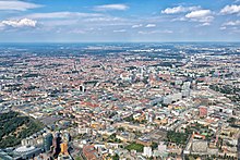 Vogelperspektive auf die Stadt Berlin, die drei Viertel der Farbfotografie einnimmt. Im unteren linken Bereich ist der Potsdamer Platz, in der Mitte des Fotos befindet sich der Fernsehturm und in der Ferne sind die ersten Felder und Wälder.