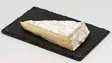 Triangle de fromage à croûte blanche d'intérieur jaune présenté sur un ardoise d'une vingtaine de centimètre de long. Le fromage fait les deux tiers de l’ardoise en longueur. Le tout est sur un fond blanc.