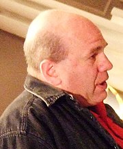 A bald man wearing a black denim jacket and red shirt looking sideways and opening his mouth to speak.