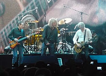 A colour photograph of John Paul Jones, Robert Plant and Jimmy Page performing on stage, with Jason Bonham partially visible on drums in the background