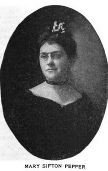 A white woman wearing eyeglasses, a black velvet choker band, and a black dress with a wide sweetheart neckline. Her hair is in an updo.