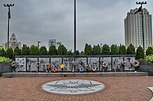 Ten large panels made of a dark stone are engraved with images from the Vietnam War. Flowers and wreaths lean against them. In front of the panels is a brick plaza with a liberty bell design.