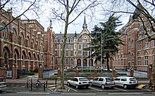 Photographie en couleur représentant trois voitures stationnées en épi au premier plan et, devant elles, une grille et un porche d'entrée donnant sur une cour d'honneur plantée d'un grand arbre à droite et encadrée de hauts bâtiments néo-gothiques dont, au centre, un porche flanqué de deux tourelles surmonté d'un campanile