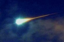 A colour photograph of a bright white ball with a firey trail against dark blue sky - a bolide or bright fireball class meteor.