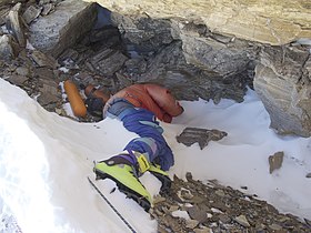 Photographie couleur en plongée d'un homme allongé entre neige et roche et au visage caché derrière son bras, emmitouflé dans d'épais vêtements et portant des chaussures d'alpinisme vertes.