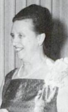 A smiling older white woman, with dark hair in a bouffant updo, wearing a corsage and pearls and a gown with a square neckline