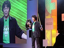 Shigeru Miyamoto standing on a stage holding replicas of Link's sword and shield
