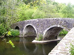 Photo d'un pont en pierres grises à trois arches enjambant une rivière bordée d'arbres feuillus