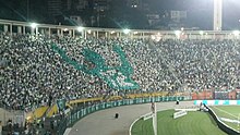Arquibancada de um estádio de futebol lotada, com a maioria usando camisas brancas, e um mosaico em verde de um jogador apontando os dedos indicadores para o céu e com a camisa 12.