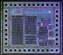 A microscope image of an integrated circuit die used to control LCDs. The pinouts are the dark circles surrounding the integrated circuit. NXP PCF8577C LCD driver with I2C (Colour Corrected).jpg