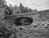 A black-white photo of a stone, arch bridge spanning a creek.