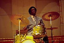 A man sit down behind a golden-colored drum kit. The man holds a pair of drumsticks and he looks concentrated while beating the cymbal at his right and the snare at the center.