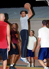 Obama holding a basketball above his head in midair while four other players look at him. He looks toward the camera over his right shoulder.