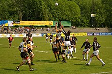 Fotografia kolorowa. Zdjęcie boiska na którym rozgrywany jest mecz rugby union pomiędzy drużyną gospodarzy, Budowlanych Łódź, w biało-czarnych strojach, a gośćmi, drużyną Folc AZS AWF Warszawa w strojach czarno-białych. Gracze obu drużyn przygotowują się do chwytu piłki z powietrza (opadającej z góry) po wykopie – gracz gospodarzy, z numerem 5, jest wynoszony do góry przez dwóch partnerów z drużyny, podczas gdy gracz gości, z numerem 11, próbuje, skacząc, samodzielnie dosięgnąć, znajdującej się powyżej, obu piłki. W tle niskie trybuny w niewielkimi grupkami kibiców, a za nimi drzewa w parku.