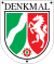 Schildförmige Denkmalplakette des Landes Nordrhein-Westfalen mit Wappen des Landes Nordrhein-Westfalen, darüber in Großbuchstaben der Schriftzug „Denkmal“, oben links und rechts sowie unten mittig ein Nagel
