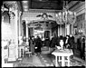 Interior de una casa con gente reunida vistiendo abrigos de invierno. Se ven adornos como candelabros y alfombras lujosas.