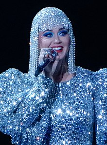 Katy Perry, a woman wearing a jeweled wig, smiling on stage wearing a jeweled outfit. She is holding a microphone to her mouth in her right hand.