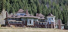 An elaborate stone and wood building with many peaked roofs and chimneys with a wooded hill behind it.