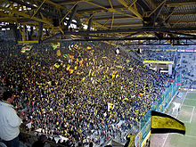 Vue depuis sous les toits d'une tribune à gauche pleine de supporters debout avec des drapeaux jaunes en plein match.
