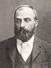 Heavily bearded man with dark receding hair, wearing a dark coloured jacket, white collar and pale tie. He is looking slightly to the left, with a solemn expression