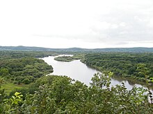 Photographie depuis une colline d'une rivière assez large dans une plaine plate recouverte de forêts.