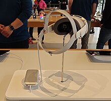 Photo of an Apple Vision Pro, on a stand, with a charger. A man stands in the background in a blue shirt.