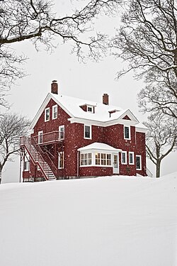 A two-story red house with a gabled roof pierced by two tall brick chimneys on the ends, and a large staircase going up the side, seen from its front left corner. Snow is falling and covering the ground