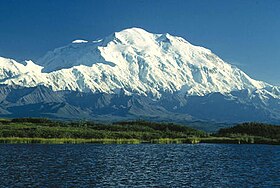 A snaw-covered, gently slopin muntain is in the backgrund, wi a loch in the foregrund