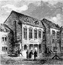 A three-story building with three men standing in front having a conversation, and one or more other people near the building.