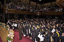 Young adults wearing ceremonial black robes and graduation caps stand at their seats in an ornate hall while onlookers in a three sided balcony applaud above them.