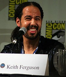 An African-American man gives a wide smile. He sitting in front of a microphone and his eyes are squinted.