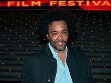 A dark skinned man with a black afro. The man is smiling and is wearing a dark green button shirt wide open on top of a buttoned up turquoise shirt.