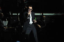 A man wearing a black suit, pants, and sunglasses is holding a microphone on his right hand