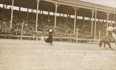 Woman in black dress roping a horse and rider in front of a grandstand.. Miss La Due Champion Lady Fancy Roper of the World at the Stampede. Calgary, Alta. 1912.