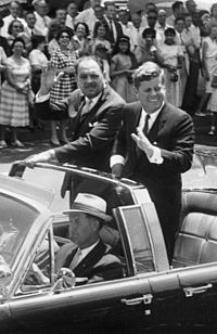 (L–R) English: Motorcade for President Mohammad Ayub Khan of Pakistan. In open car (Lincoln-Mercury Continental with bubble top): Secret Service agent William Greer (driving); Military Aide to the President General Chester V. Clifton (front seat, centre); Secret Service Agent Gerald "Jerry" Behn (front seat, right, partially hidden); President Mohammad Ayub Khan (standing); President John F. Kennedy (standing). Crowd watching. 14th Street, Washington, D.C.