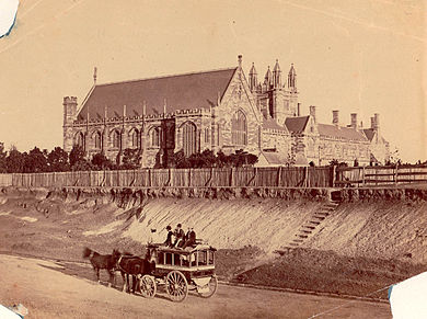 An old sepia photograph shows Sydney University from the west. Only the front section and Great Hall of the Main Building exists. It is separated from the road by a paling fence and a steep eroded incline, where there is now a garden. A horse-drawn bus with passengers up-top is standing in the road.