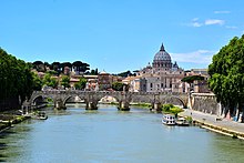 A view of Rome on a sunny afternoon looking along the river. A bridge crosses the river and beyond it is a hill on which the grey dome of St. Peter's rises above ancient buildings and dark pine trees.