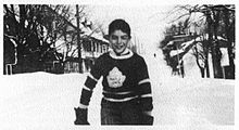 Мальчик стоит на заснеженной улице. На нем темный свитер со стилизованным логотипом в виде кленового листа на груди.