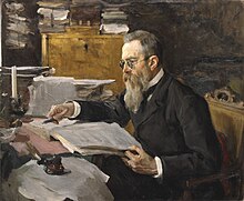 A man with dark gray hair, glasses and a long beard seated at a desk, looking at a music manuscript