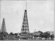 Photograph of Westinghouse "Old No.1" natural gas derrick in daytime, sometime between June 1884 and end of 1889, located at the Solitude estate in present-day Westinghouse Park. The Westinghouse carriage house/laboratory and the mansion are seen on the right.[9]