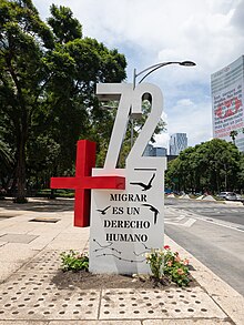 A red plus sign and the white digits "72" on a white pedestal with the Spanish words "Migrar es un derecho humano" in black uppercase letters
