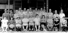 Photo noir et blanc d'un groupe assis devant un bâtiment. La plupart sont des hommes en uniformes, avec quelques femmes.