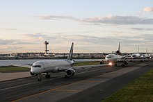 Пять гигантских самолетов стоят в очереди на взлетно-посадочной полосе рядом с небольшим водоемом. Позади них вдали находится аэропорт и диспетчерская. 